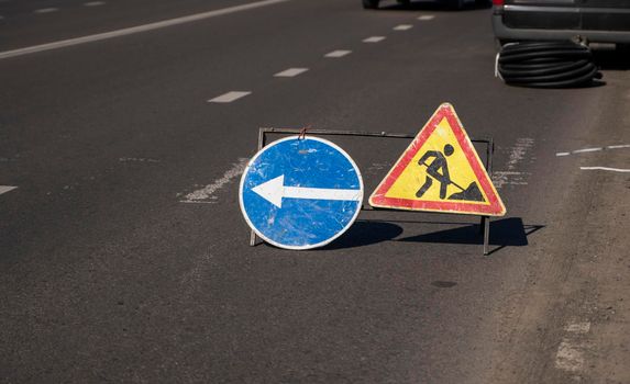 Road works sign on the road. Repair of road signs and a bypass arrow against the background of a torn road. Repair work in the middle of the carriageway, selective focus