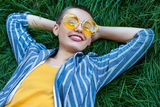 Portrait of Happy fancy young woman with short hair in casual blue striped suit, yellow shirt, glasses lying down on green grass, holding hands behind head, looking with toothy smile. summertime shot