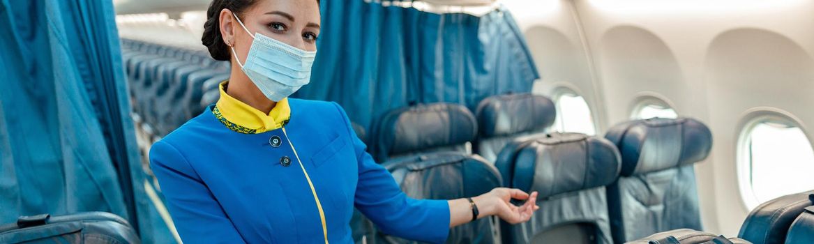 Female flight attendant in protective face mask welcoming on board of airplane while gesturing towards passenger chair