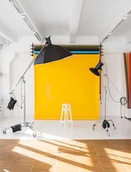 Yellow background roll with a white chair and two professional flash on a c-stands. Sunny daylight photo studio interior with set of professional equipment