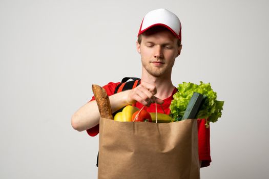 Grocery delivery courier man in a red cap and t-shirt with a thermal bag on a shoulders holds paper bag with food on white studio background. Food delivery service, online shopping