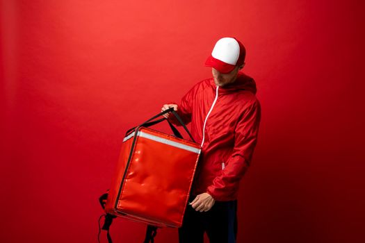 Professional delivery employee man 20s in red cap T-shirt uniform workwear work as dealer courier with red thermal food bag backpack isolated on red background. Service concept