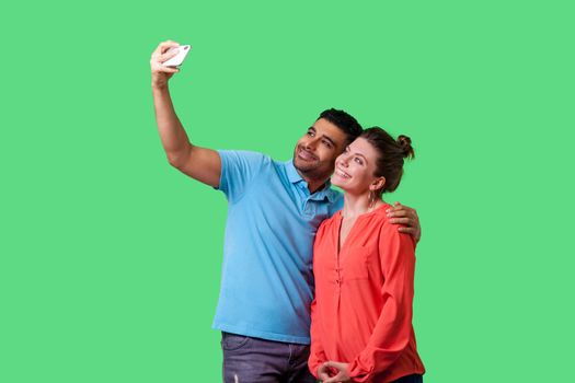 Couple doing selfie. Portrait of positive young man and woman in casual wear standing, taking pictures together using cellphone, happy memories. isolated on green background, indoor studio shot