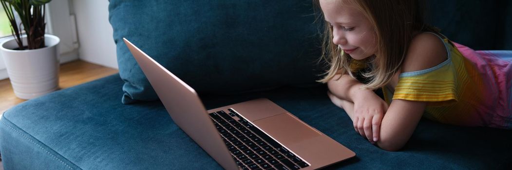 A little girl is lying on the couch with a laptop, close-up. The child participates in a video conference, remote communication with parents