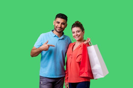Satisfied shoppers. Portrait of happy bearded young man in casual clothes pointing at shopping bags in hands of excited woman, boasting discounts. isolated on green background, indoor studio shot