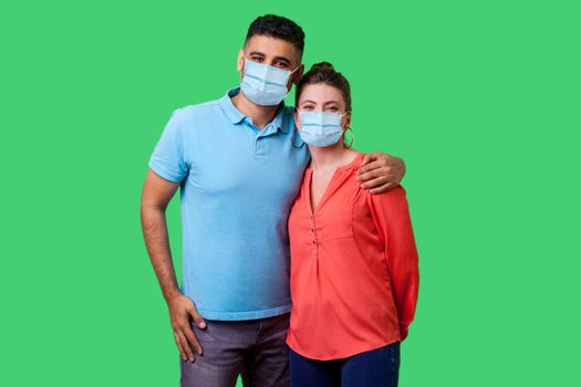 Portrait of young attractive family couple with surgical medical mask standing together, embracing and looking at camera with smile, strong relations. isolated on green background, indoor studio shot