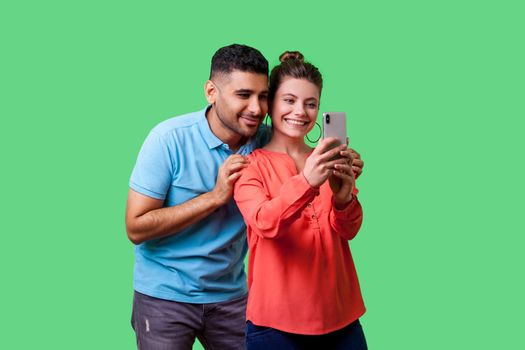 Couple in love taking picture together. Portrait of happy beautiful woman with charming toothy smile in casual clothes doing selfie with boyfriend. isolated on green background, indoor studio shot