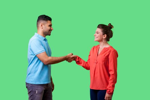 Portrait of positive friendly man and woman in casual wear standing, smiling at each other and shaking hands, couple first meeting or acquaintance. isolated on green background, indoor studio shot