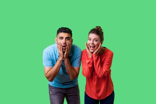 Wow. Portrait of shocked attractive young couple in casual wear standing together, holding hands on face with surprised amazed expression, open mouth. isolated on green background, indoor studio shot