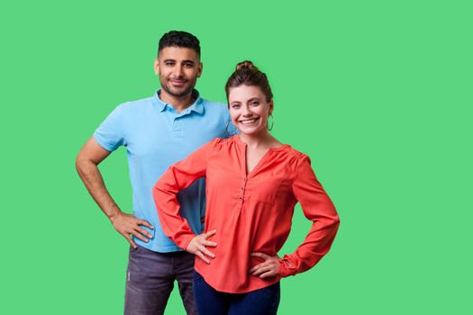 Portrait of confident attractive young couple in casual clothing standing together, holding hands on hips, smiling at camera, looking proud and happy. isolated on green background, indoor studio shot