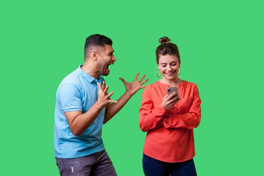 Furious man in casual wear standing with raised arms and desperately screaming near happy woman using phone and ignoring him, attracting her attention. isolated on green background, indoor studio shot