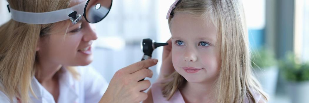 A woman otolaryngologist in a clinic looks at the ear of a little girl. Children's doctor, diagnosis of diseases, consultation