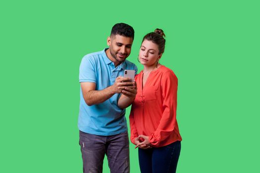 Portrait of curious attractive young couple in casual wear standing, reading message together on mobile phone, looking concentrated and interested. isolated on green background, indoor studio shot