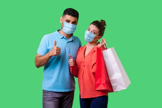 Portrait of satisfied young couple with surgical medical masks showing thumbs up gesture together, cute woman holding bags and smiling. isolated on green background, indoor studio shot