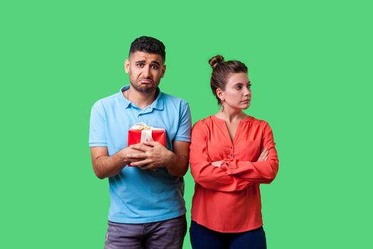 Portrait of angry resentful woman in casual clothes standing with crossed arms, disappointed with gift, turned away from man holding bad present. isolated on green background, indoor studio shot