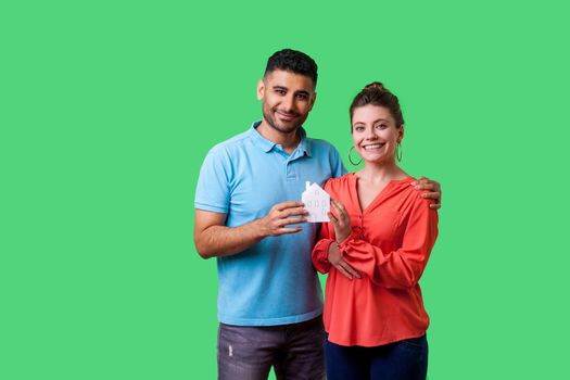 Portrait of lucky young couple in casual clothes looking at camera with toothy smile, both holding paper house, satisfied with home purchase, mortgage. isolated on green background, indoor studio shot