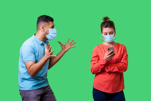 Furious man with surgical medical mask standing with raised arms and desperately screaming near woman using phone and ignoring him, attracting her attention. isolated on green background, indoor shot