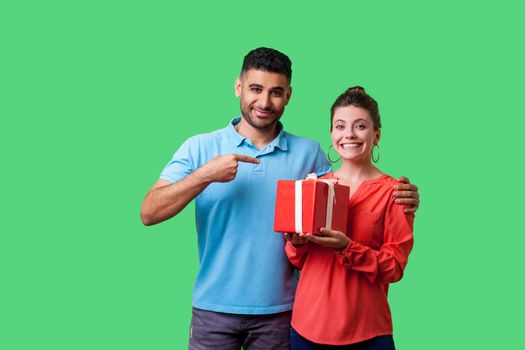 Best present for her. Portrait of happy couple in casual wear, man pointing at excited woman with gift box, looking satisfied and smiling at camera. isolated on green background, indoor studio shot