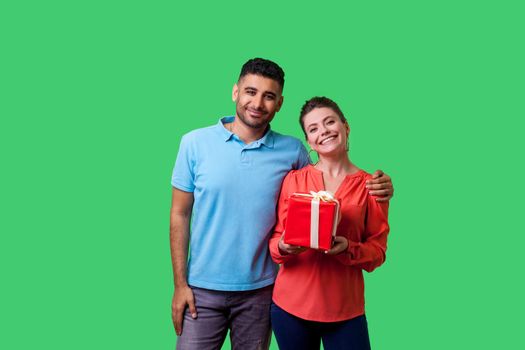 Portrait of satisfied young couple in casual wear hugging, delighted woman holding gift box, holiday present, looking at camera with toothy smile. isolated on green background, indoor studio shot