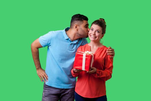 Family celebrating Valentines day. Young man in casual clothes kissing woman on cheek, she's holding gift box and looking up dreamy and surprised. isolated on green background, indoor studio shot