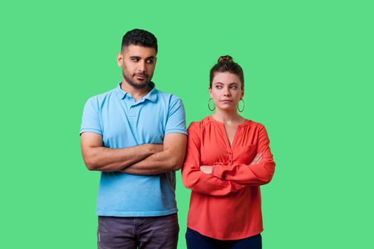 Portrait of upset young couple in casual wear standing together with crossed hands, looking sideways at each other with resentful glance, suspicion. isolated on green background, indoor studio shot