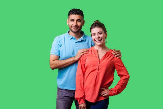 Portrait of happy attractive young couple in casual wear standing together, looking at camera with toothy smile, caring man holding female shoulders. isolated on green background, indoor studio shot