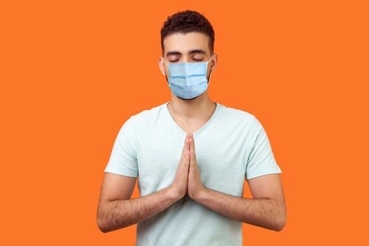 Yoga practice for mind balance. Portrait of peaceful man with surgical medical mask in white t-shirt holding hands in namaste or prayer, meditating. indoor studio shot isolated on orange background