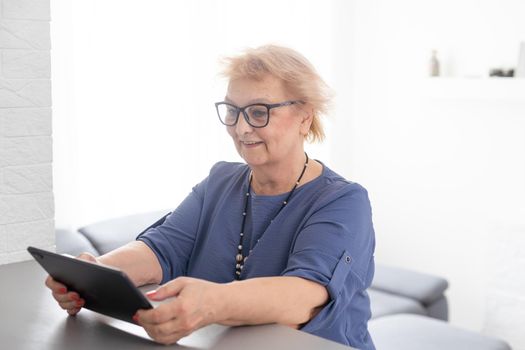Merry mature middle-aged woman spends leisure time at home using a digital tablet. Cheerful modern senior lady holds computer, watching funny movies and laughs. Leisure and technology concept