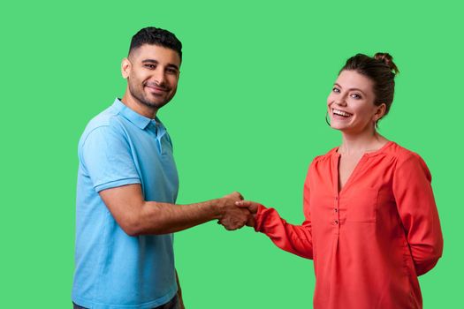 Portrait of lovely young couple in casual wear standing, looking at camera with smile and shaking hands, friendly meeting or acquaintance, greeting. isolated on green background, indoor studio shot