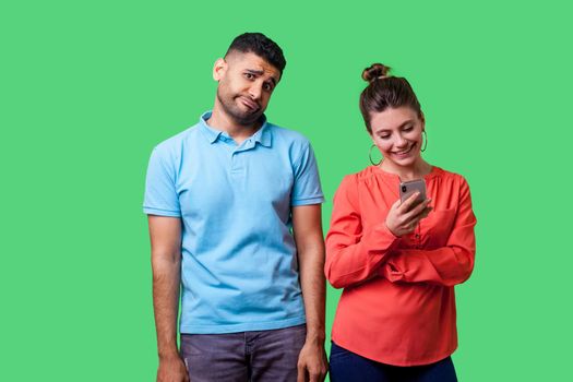 Portrait of funny upset man in casual wear standing with hands down, looking bored and annoyed, upset about woman using smartphone, ignoring him. isolated on green background, indoor studio shot