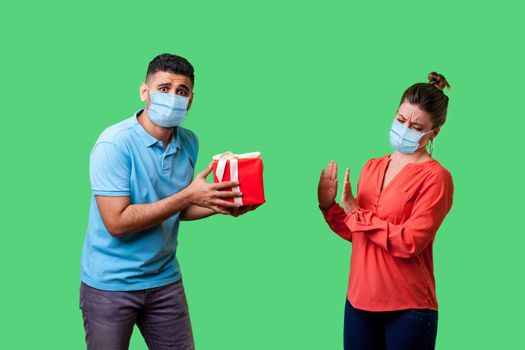 Portrait of upset resentful man with surgical medical mask giving present to naughty woman, dissatisfied girl showing refusal gesture, bad gift. isolated on green background, indoor studio shot