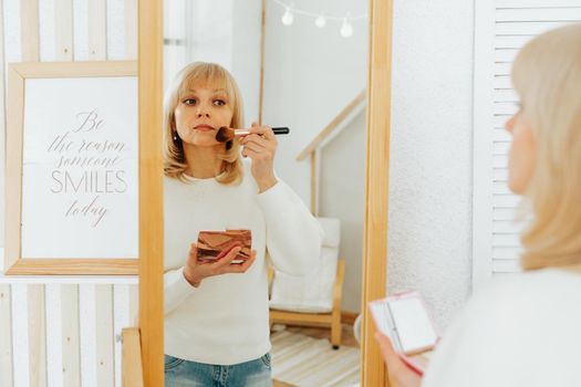 Beautiful smiling mature senior caucasian woman with blonde hair standing near mirror with make up palette and brush, doing makeup and looking at reflection in living room at home.