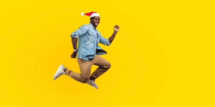 Full length portrait of happy joyous man with new year santa hat jumping or flying, hurry running to his dream, looking at camera with toothy smile. indoor studio shot isolated on yellow background