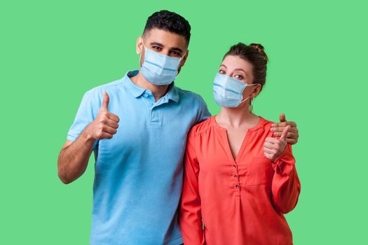 Portrait of positive young couple with surgical medical mask standing together, hugging as friends, showing thumbs up gesture and smiling at camera. isolated on green background, indoor studio shot