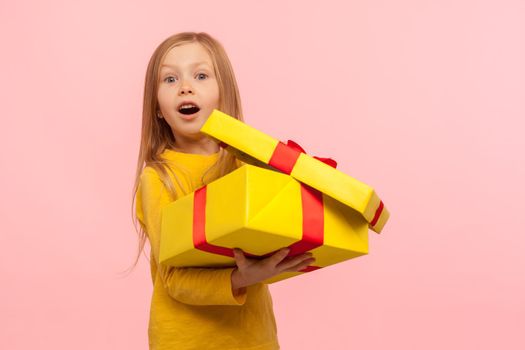 Charming child surprised by birthday present. Portrait of cute little girl opening gift box and keeping mouth open in amazement, shocked expression. indoor studio shot isolated on pink background