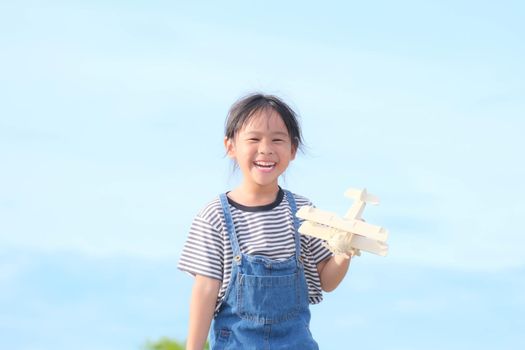 Happy kid playing with toy plane against blue summer sky background. Cute little girl running through the meadow on a sunny day with a toy plane in hand. Childhood dream imagination concept.