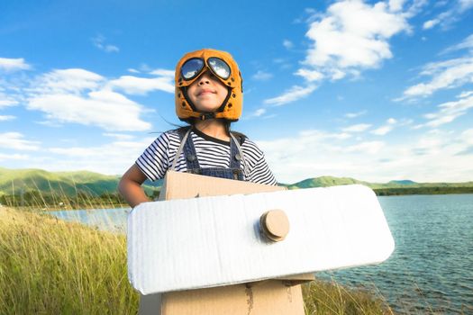 Cute dreamer little girl playing with cardboard planes in a lakeside meadow against blue summer sky background on a sunny day. Childhood dream imagination concept.