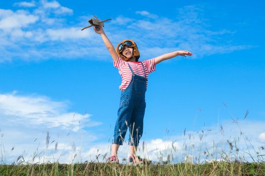 Cute little girl running through the meadow on a sunny day with a toy plane in hand. Happy kid playing with cardboard plane against blue summer sky background. Childhood dream imagination concept.