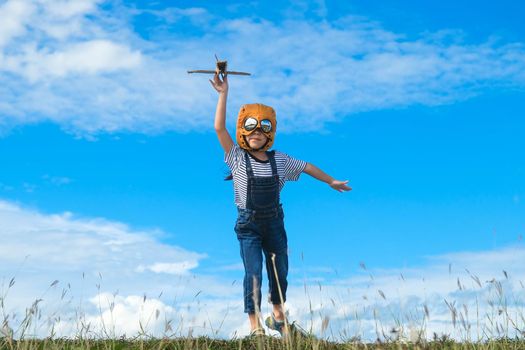 Cute little girl running through the meadow on a sunny day with a toy plane in hand. Happy kid playing with cardboard plane against blue summer sky background. Childhood dream imagination concept.