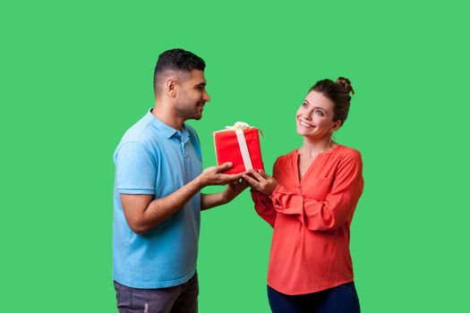 Couple celebrating Valentines day. Young handsome man in casual clothes giving box with present to beautiful elegant woman, looking at her with love. isolated on green background, indoor studio shot