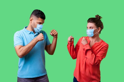 Come on let's fight. Portrait of couple with surgical medical mask standing together, keeping fists clenched and looking at each other ready to fight, joke. isolated on green background, indoor