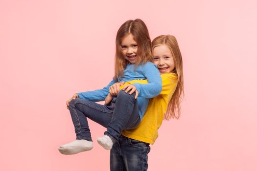 Portrait of cute preschool girl lifting up her little sister and smiling, friends having fun together, playing game, expressing carefree childish happiness. studio shot isolated on pink background