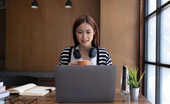 Calm and relaxed young Asian woman sipping a morning coffee while reading an online content on her portable laptop at the desk..