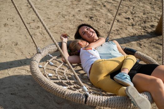 Mom and daughter swing on a round swing. Caucasian woman and little girl have fun on the playground