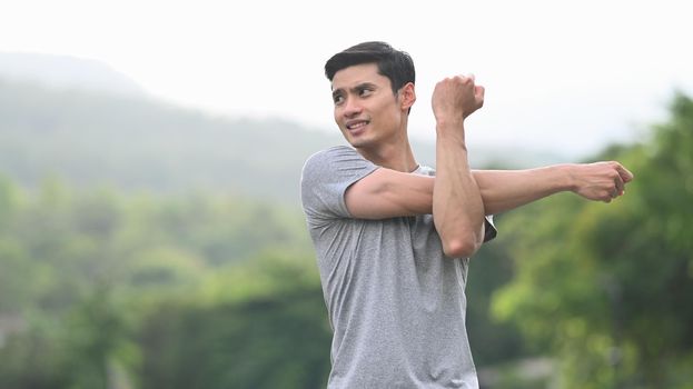 Sporty asian man stretching his arm, warming up before morning workout at the nature park. Healthy lifestyle, workout and wellness concept.
