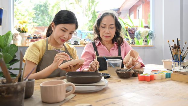 Focused young woman and mature woman painting pottery bowl in workshop. Indoors lifestyle activity, handicraft, hobbies concept.