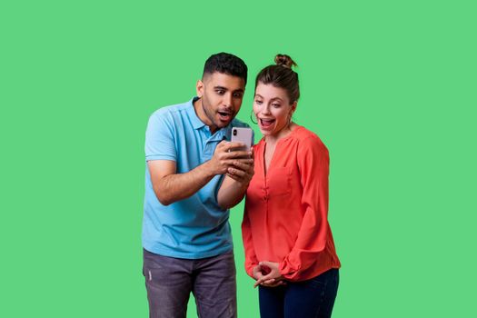 Friends reading shocking news. Portrait of amazed attractive young couple in casual wear reading unexpected message with open mouth, using mobile phone. isolated on green background indoor studio shot