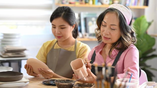 Attractive mature woman and young woman painting pottery bowl in workshop. Indoors lifestyle activity, handicraft, hobbies concept.