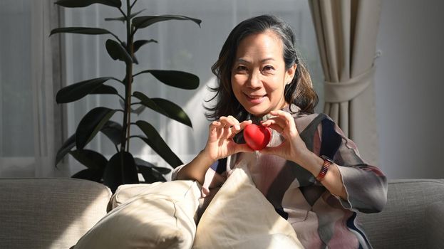 Happy middle aged woman holding heart shape. Elderly care, insurance, heart health and quality of life for elderly concept.