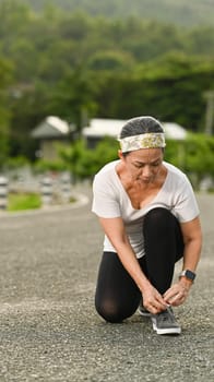 Middle aged lady in sportswear tying shoelaces before running, getting ready for jogging outdoors. Healthy lifestyle, workout and wellness concept.